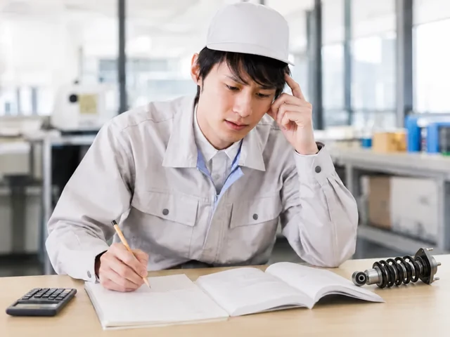 机に置いたコイルスプリング(ばね・ダンパ)を前に、ノートに書きながら考え込む若手設計者